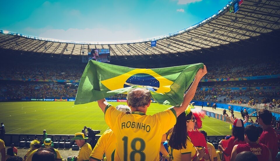 Torcedor no estádio com a camiseta amarela levantando a bandeira do Brasil em direção ao gramado