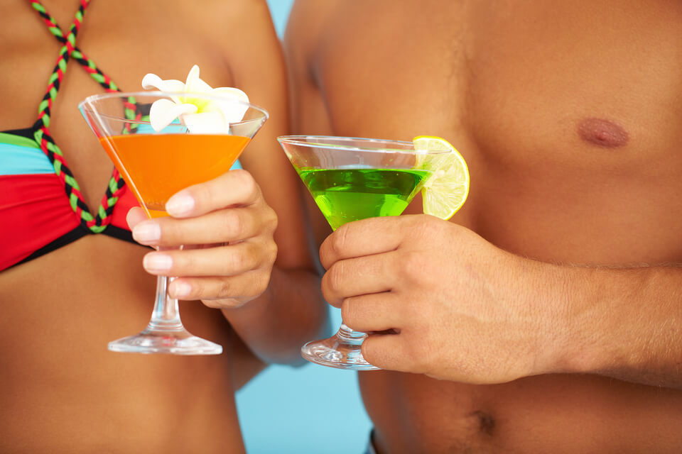 Duas pessaos de trajes de banho brindando com dois drinks.